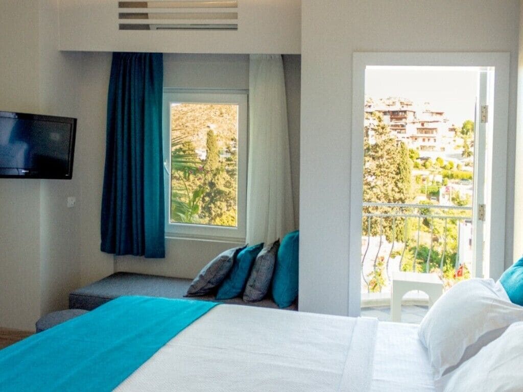 Un dormitorio con cama, televisor en la pared, ventana con cortinas y vistas a edificios.