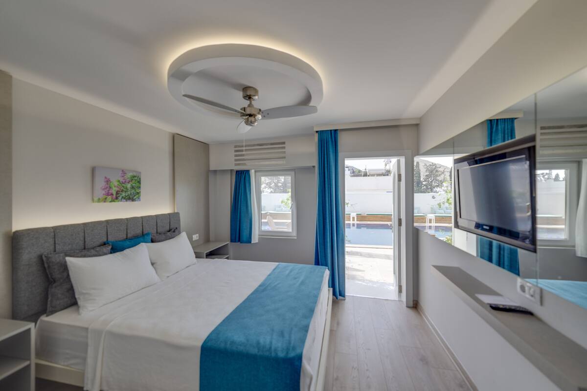 A modern hotel room with a bed, a ceiling fan, a TV, and a view of the outdoors.