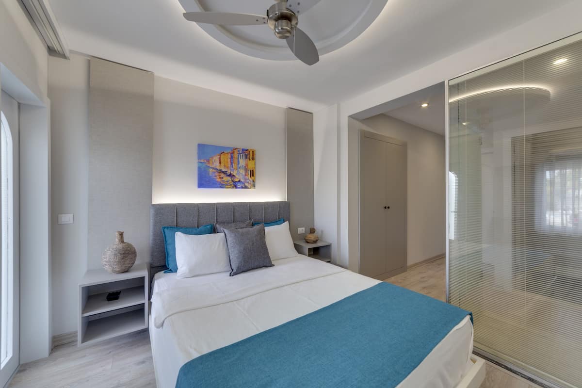 A modern hotel room with a large bed, a ceiling fan, a painting on the wall, and a glass partition.