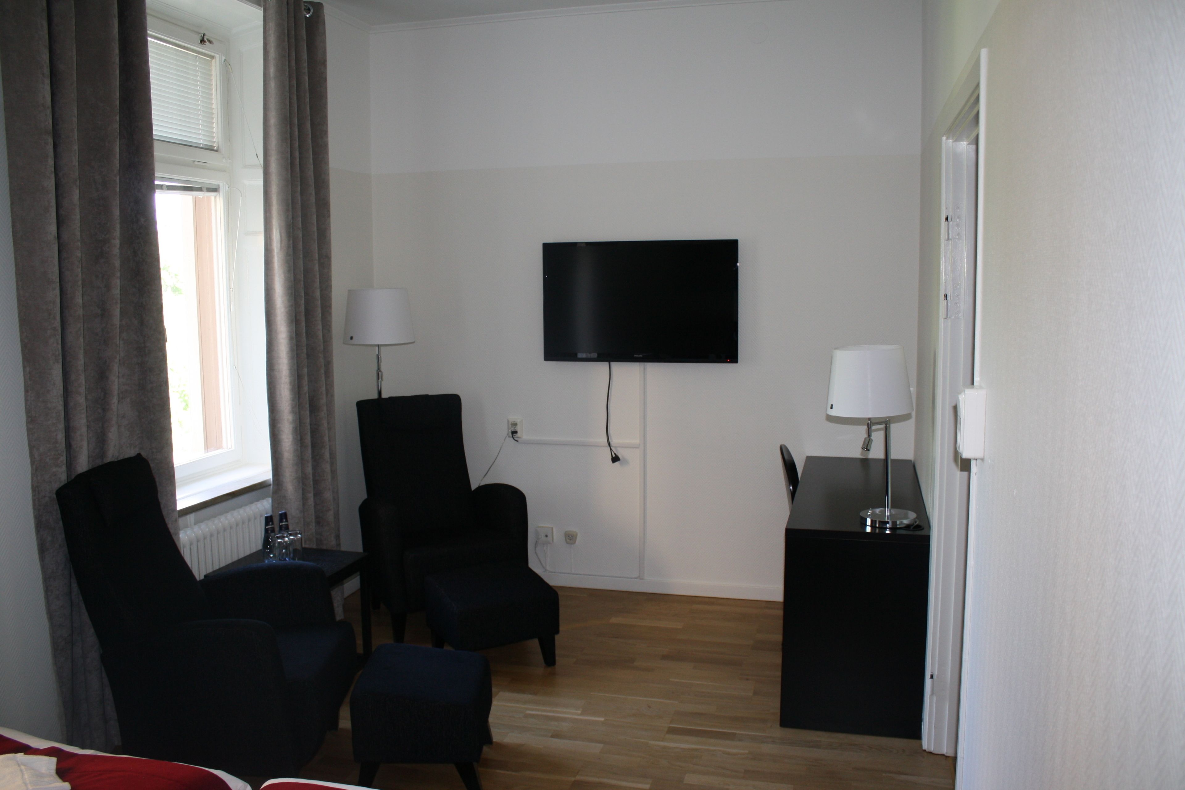 A modern hotel room with a flat-screen TV mounted on the wall, a desk with a lamp, two chairs, and a small table with bottles.