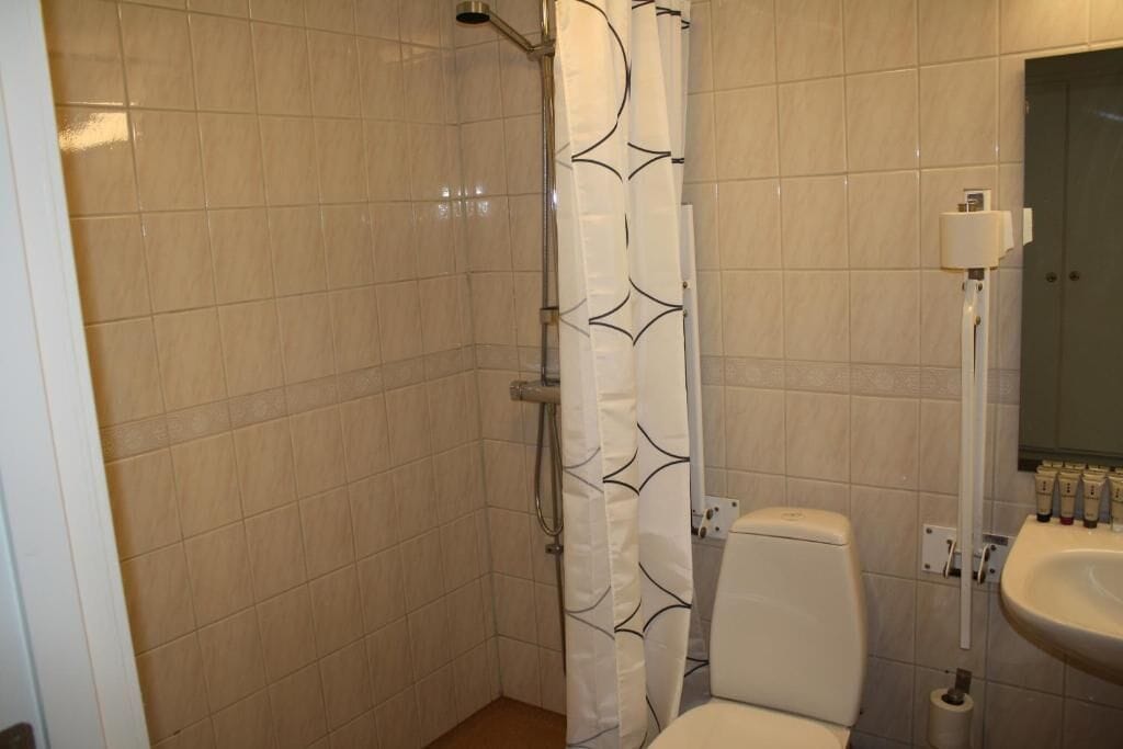 Un baño con baldosas color crema, una ducha con cortina blanca, un inodoro y un lavabo con artículos de aseo.