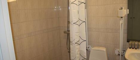 Un baño con baldosas color crema, una ducha con cortina blanca, un inodoro y un lavabo con artículos de aseo.