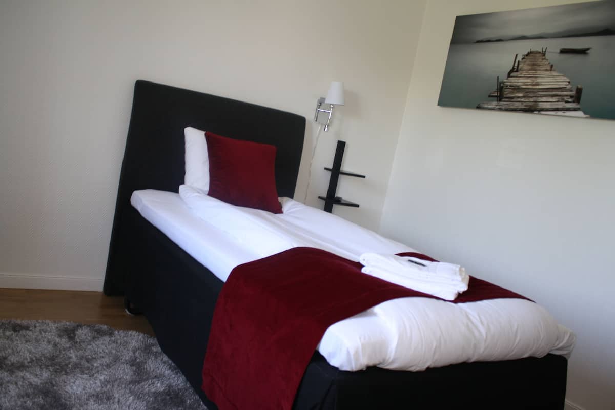 A neatly made bed with white linens and a red pillow, a black headboard, and a painting of a pier on the wall.