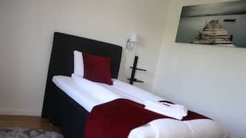 Una cama bien tendida con sábanas blancas y una almohada roja, un cabecero negro y un cuadro de un muelle en la pared.