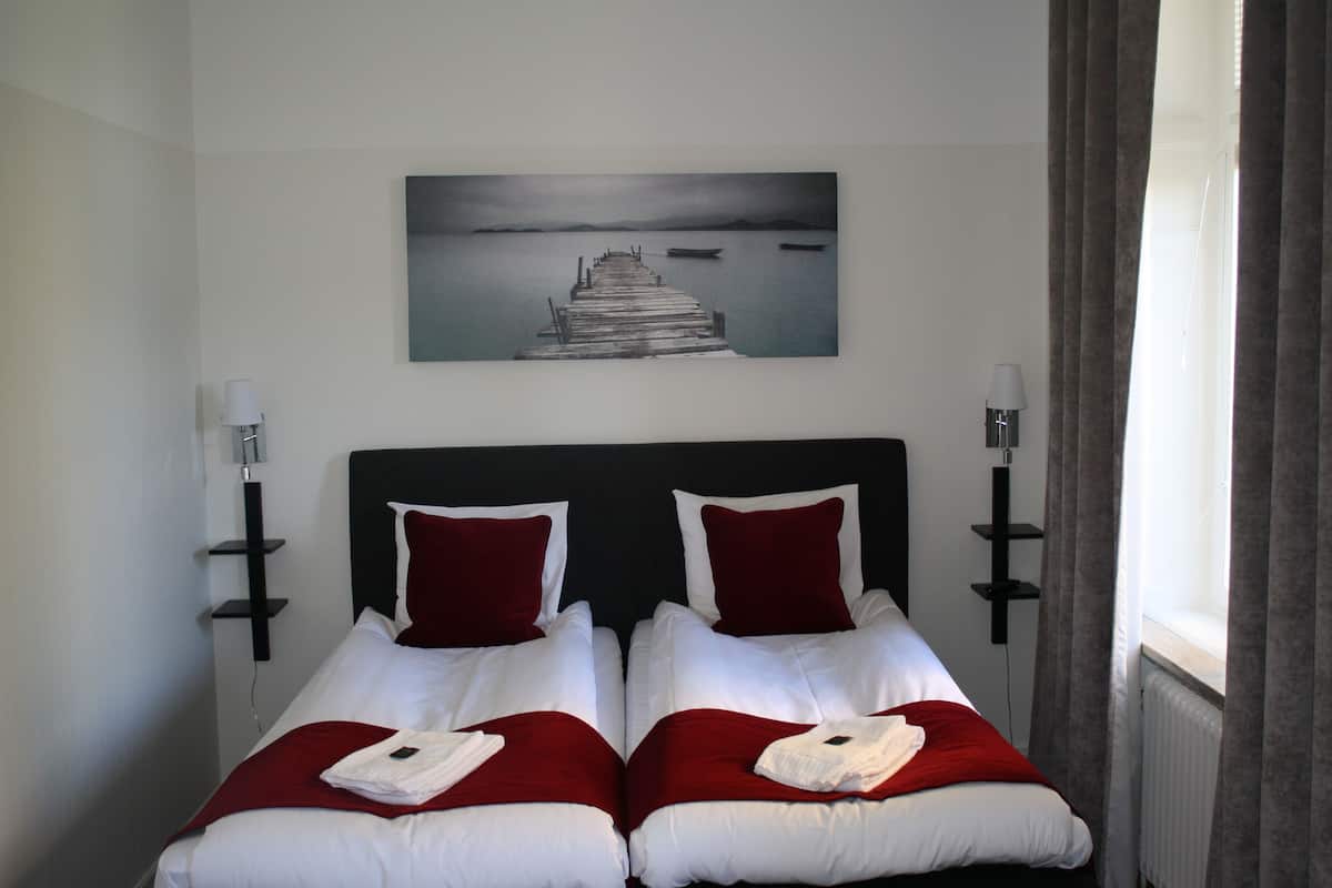 A double bed with white and red bedding, a painting of a pier on the wall, and a window with curtains.