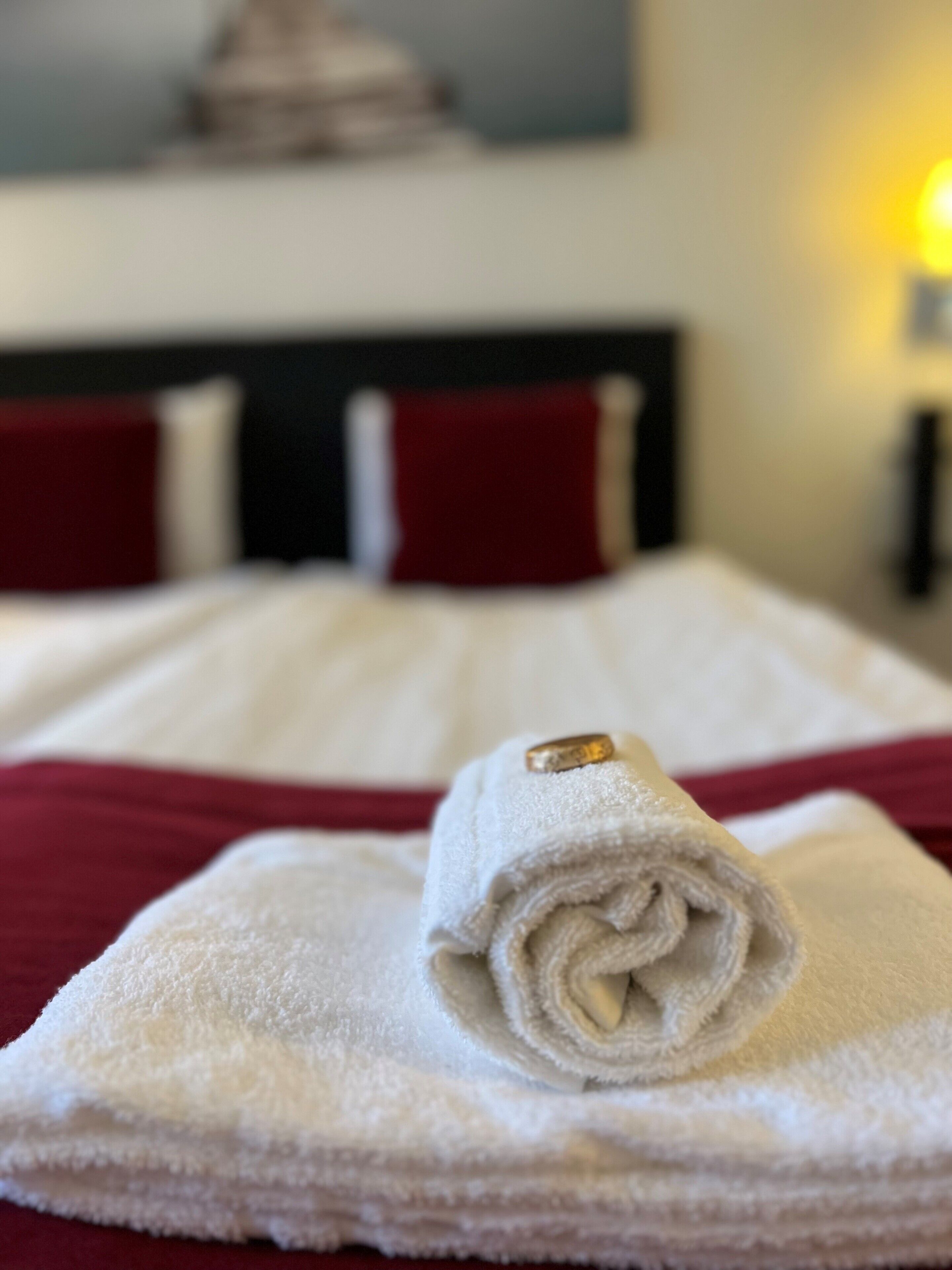 A neatly made bed with a rolled white towel and a gold ring on top, red pillows, and a lamp in the background.