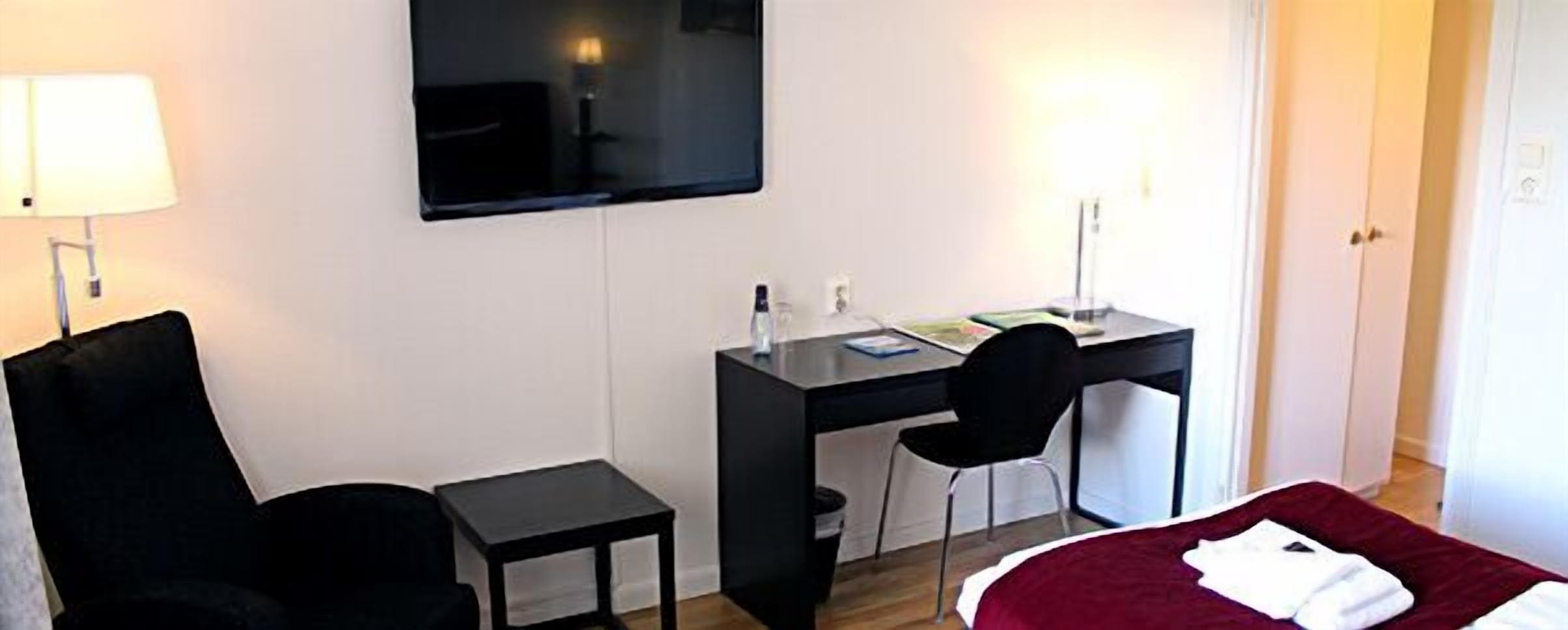 A hotel room with a bed, a desk, a chair, a small table, and a TV mounted on the wall.