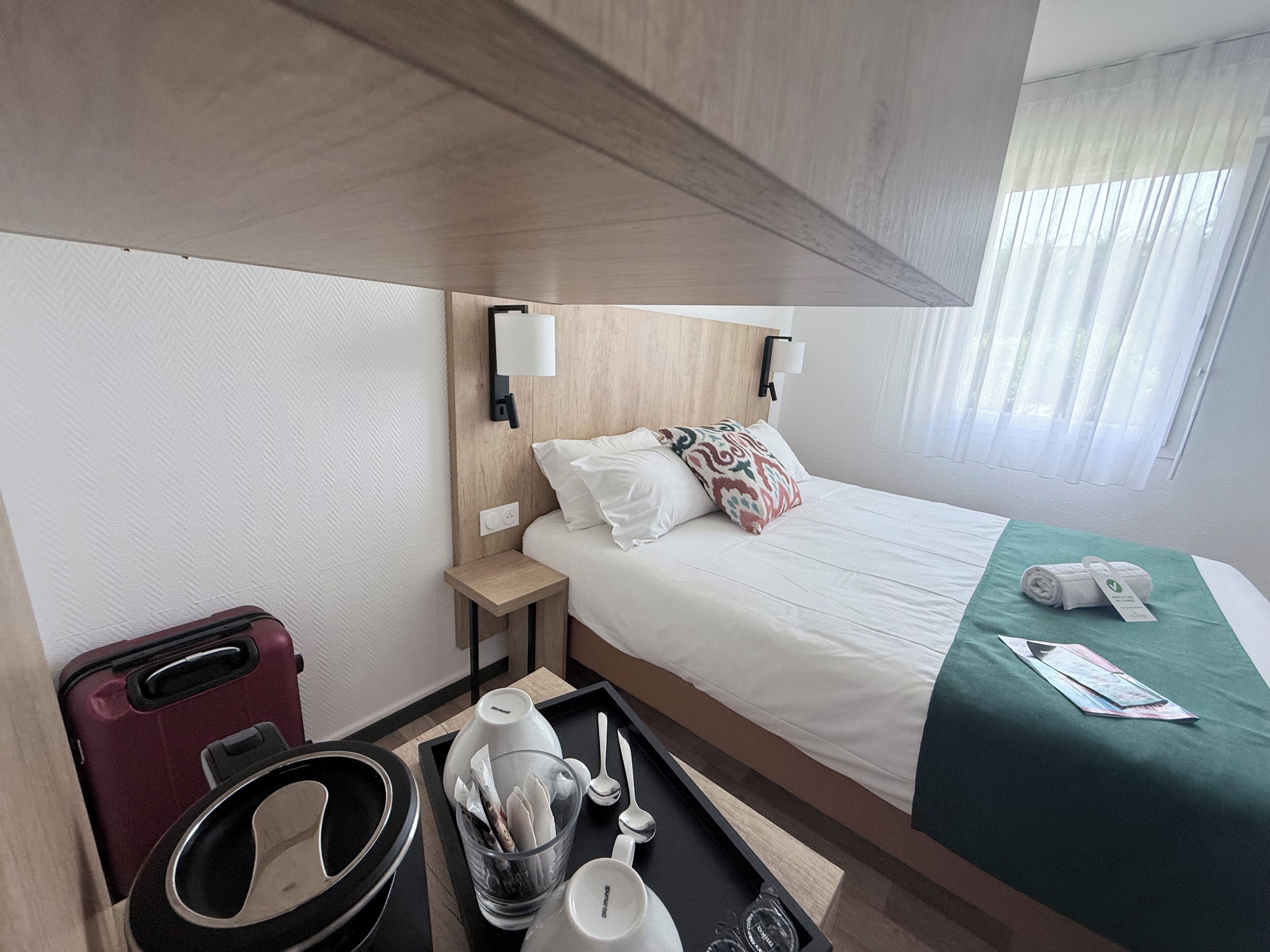 Ein Hotelzimmer mit einem Bett, einem Nachttisch, einer Lampe, einem Koffer, einem Tablett mit Pflegeartikeln und einem Fenster mit Vorhängen.