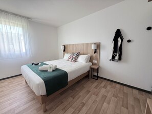 Een hotelkamer met een bed, nachtkastje, lamp, jas hangend aan de muur en een raam met gordijnen.