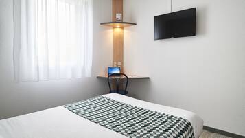 Habitación de hotel con cama, escritorio con una laptop, televisor montado en la pared y ventana con cortinas translúcidas.