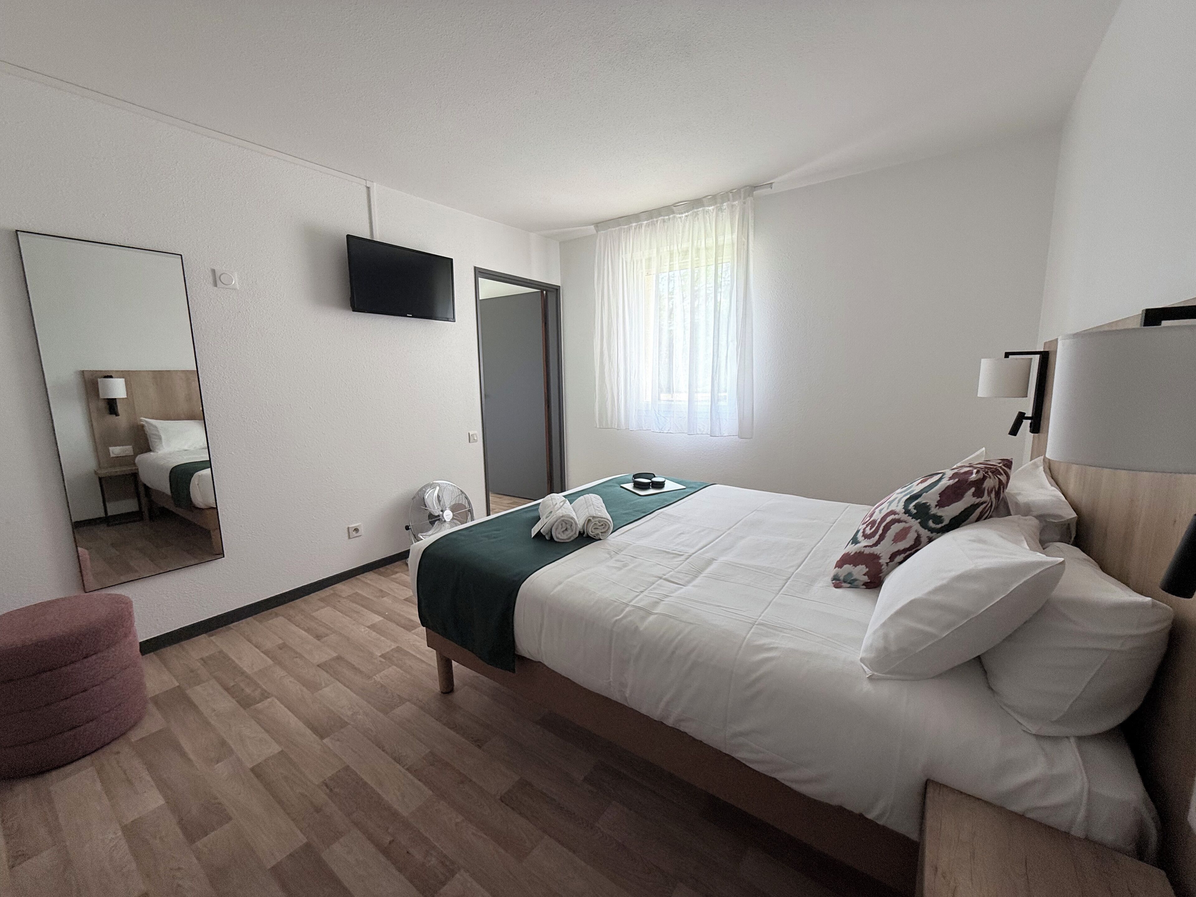 Une chambre d’hôtel avec un lit, un miroir, une télévision, une fenêtre avec des rideaux et une table de chevet.