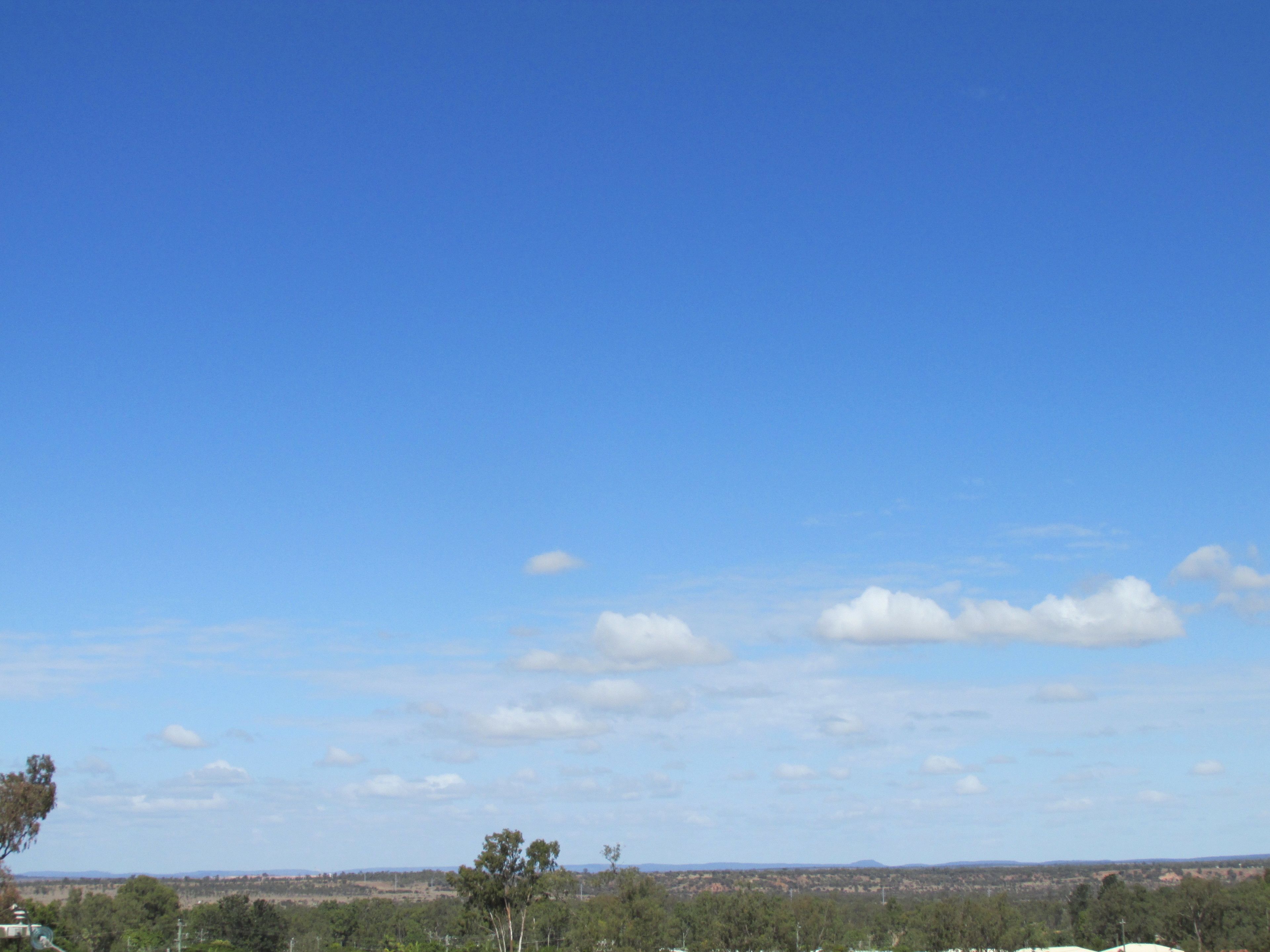 Cielo azul despejado con algunas nubes sobre un paisaje con árboles y colinas distantes.