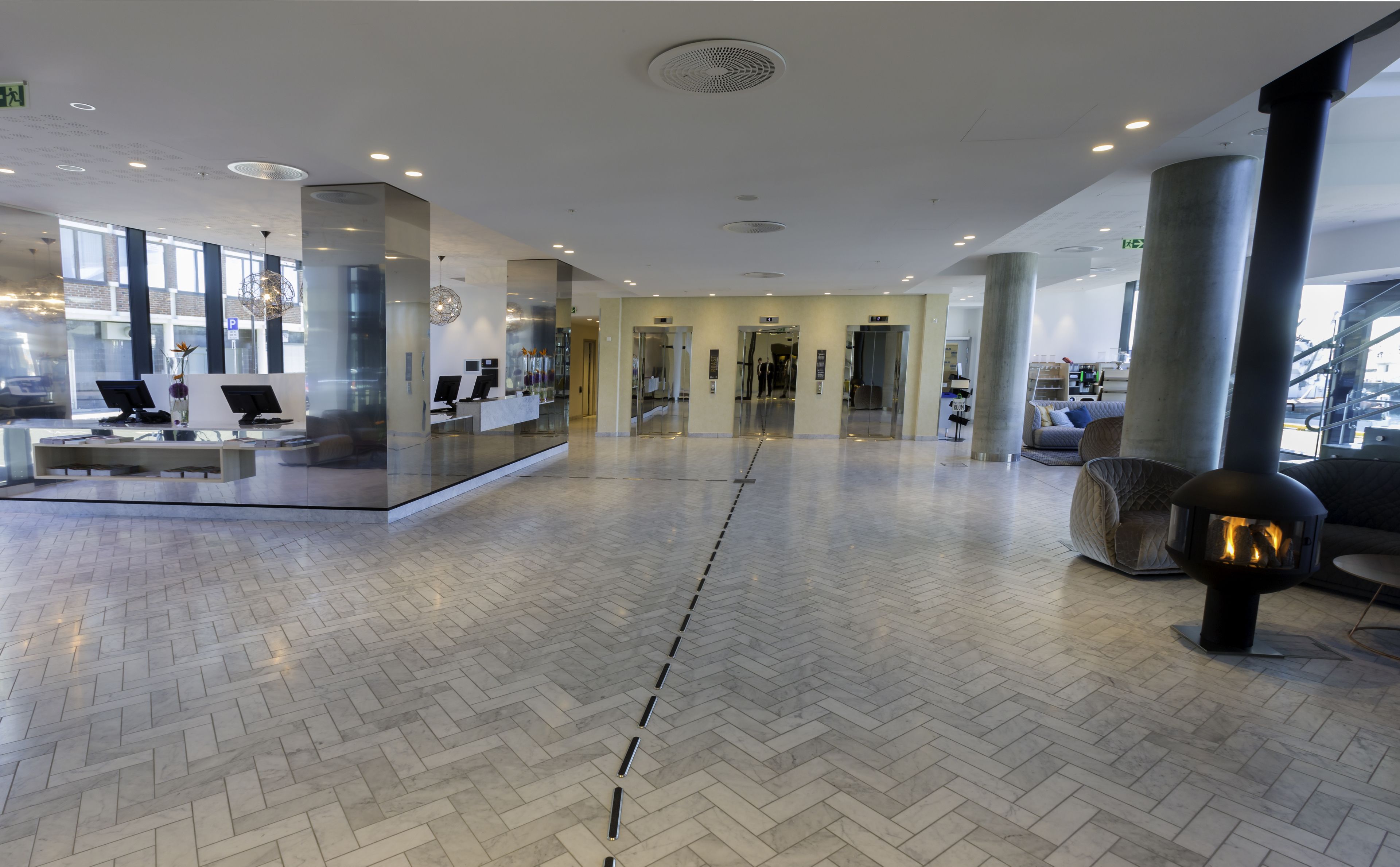 A spacious lobby with a herringbone floor pattern, a reception desk with computers, and a seating area with a modern fireplace.