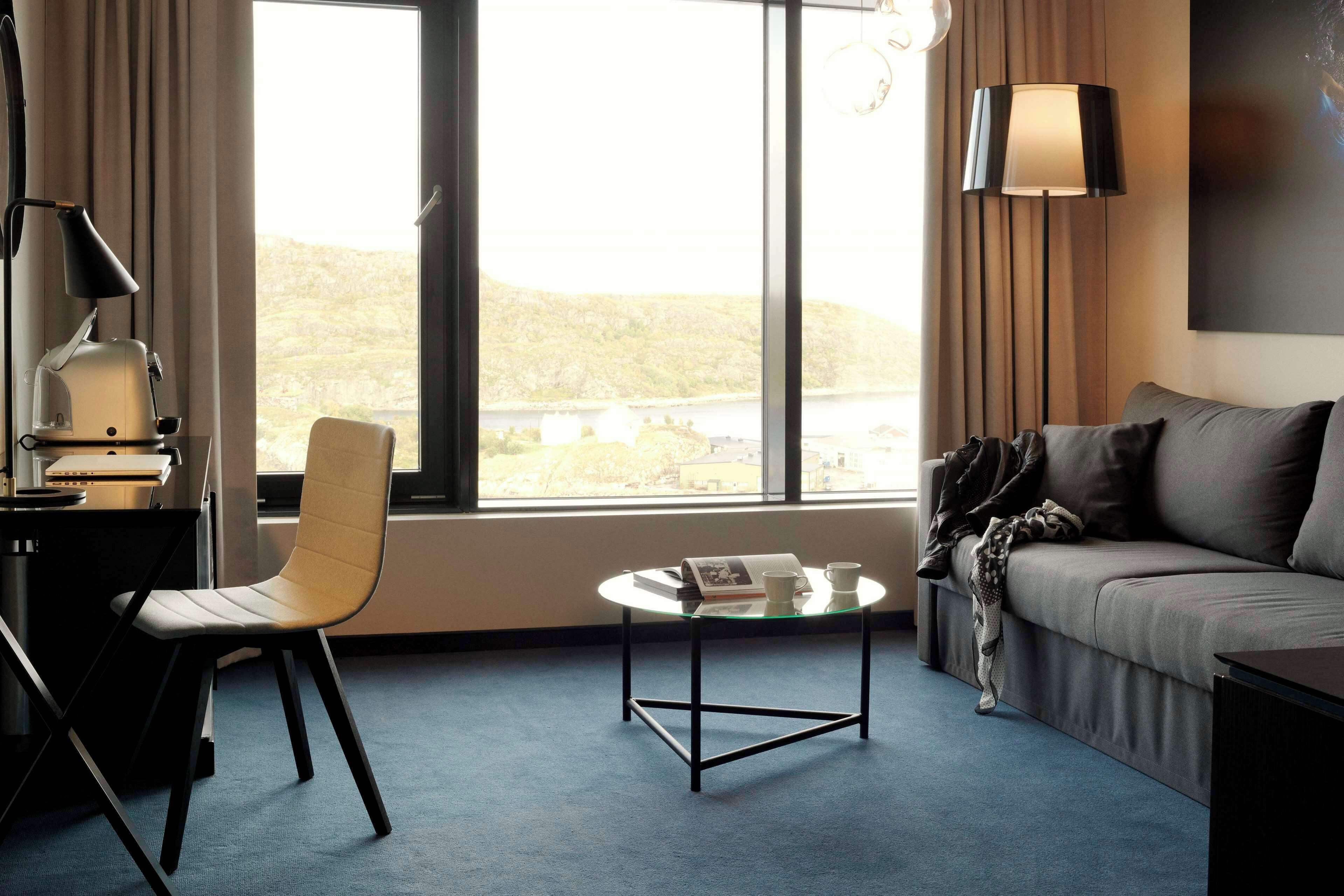 A modern hotel room with a large window, a sofa, a glass coffee table, a desk with a lamp, and a chair.