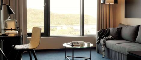 A modern hotel room with a large window, a sofa, a glass coffee table, a desk with a lamp, and a chair.