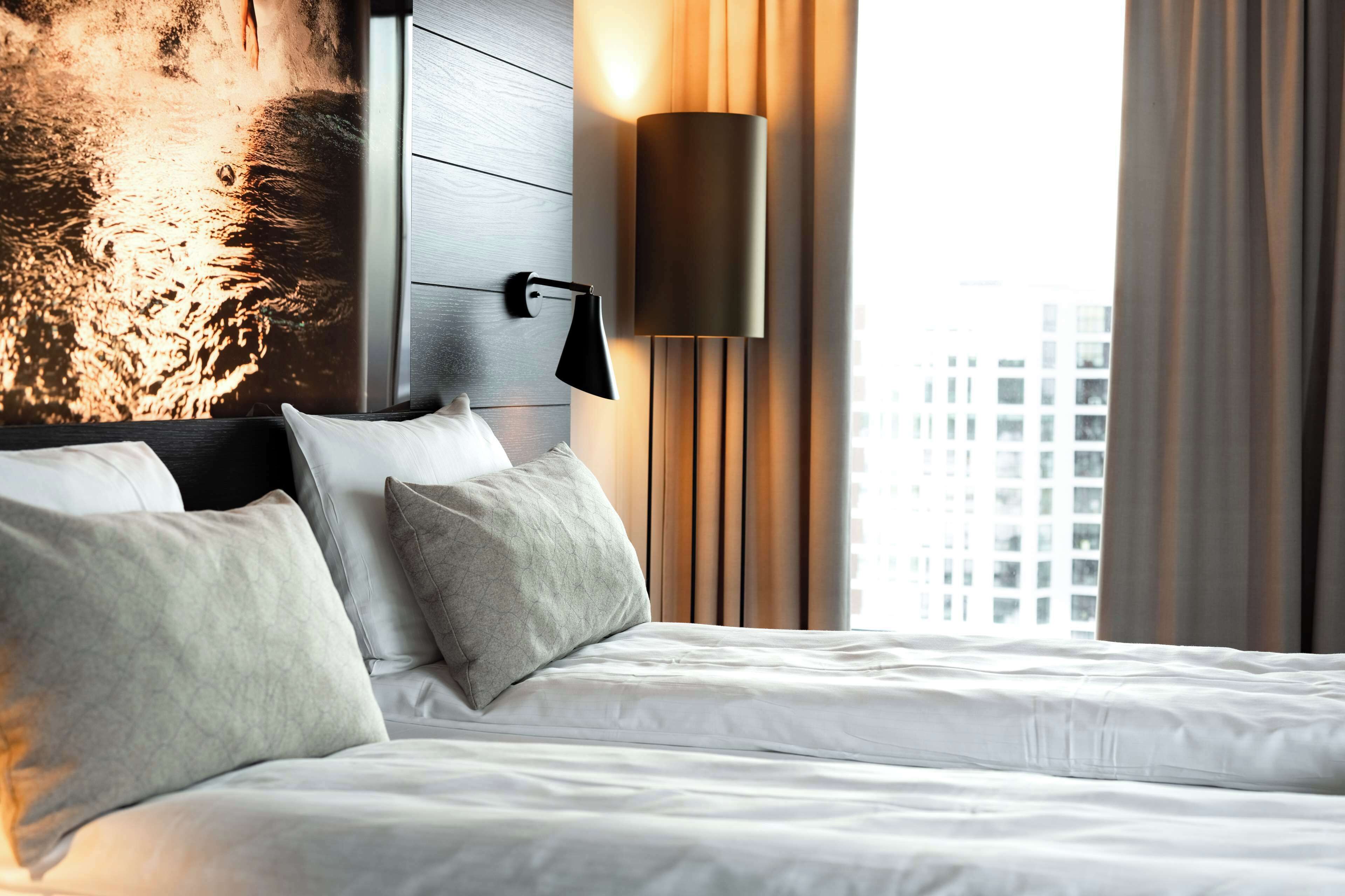 Habitación de hotel con cama, almohadas, una lámpara fijada a la pared y vistas de edificios a través de una ventana.