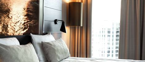 Habitación de hotel con cama, almohadas, una lámpara fijada a la pared y vistas de edificios a través de una ventana.