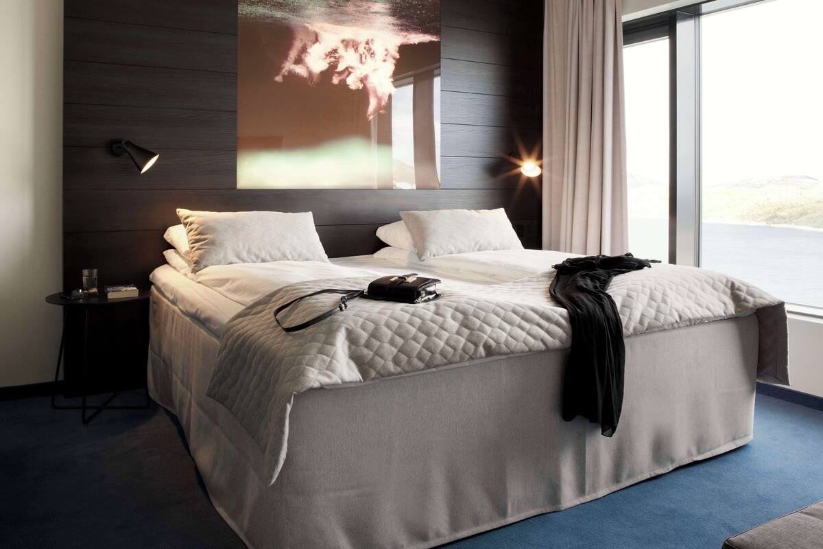 A double bed with a quilted cover, a bedside table with a lamp, and a large window with a view of the outdoors.