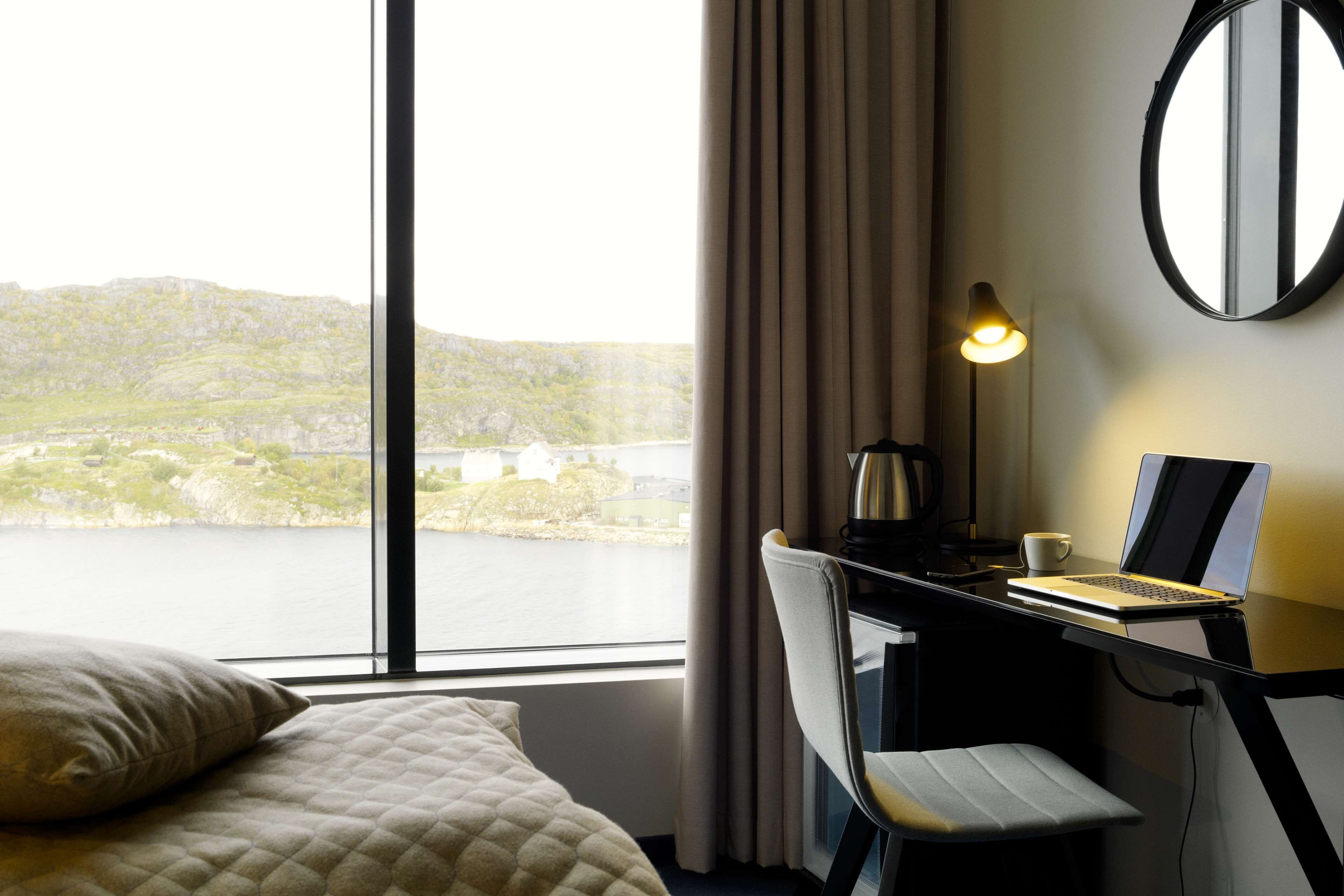 A hotel room with a bed, a desk with a laptop, a lamp, and a view of a lake and hills.