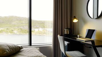 A hotel room with a bed, a desk with a laptop, a lamp, and a view of a lake and hills.