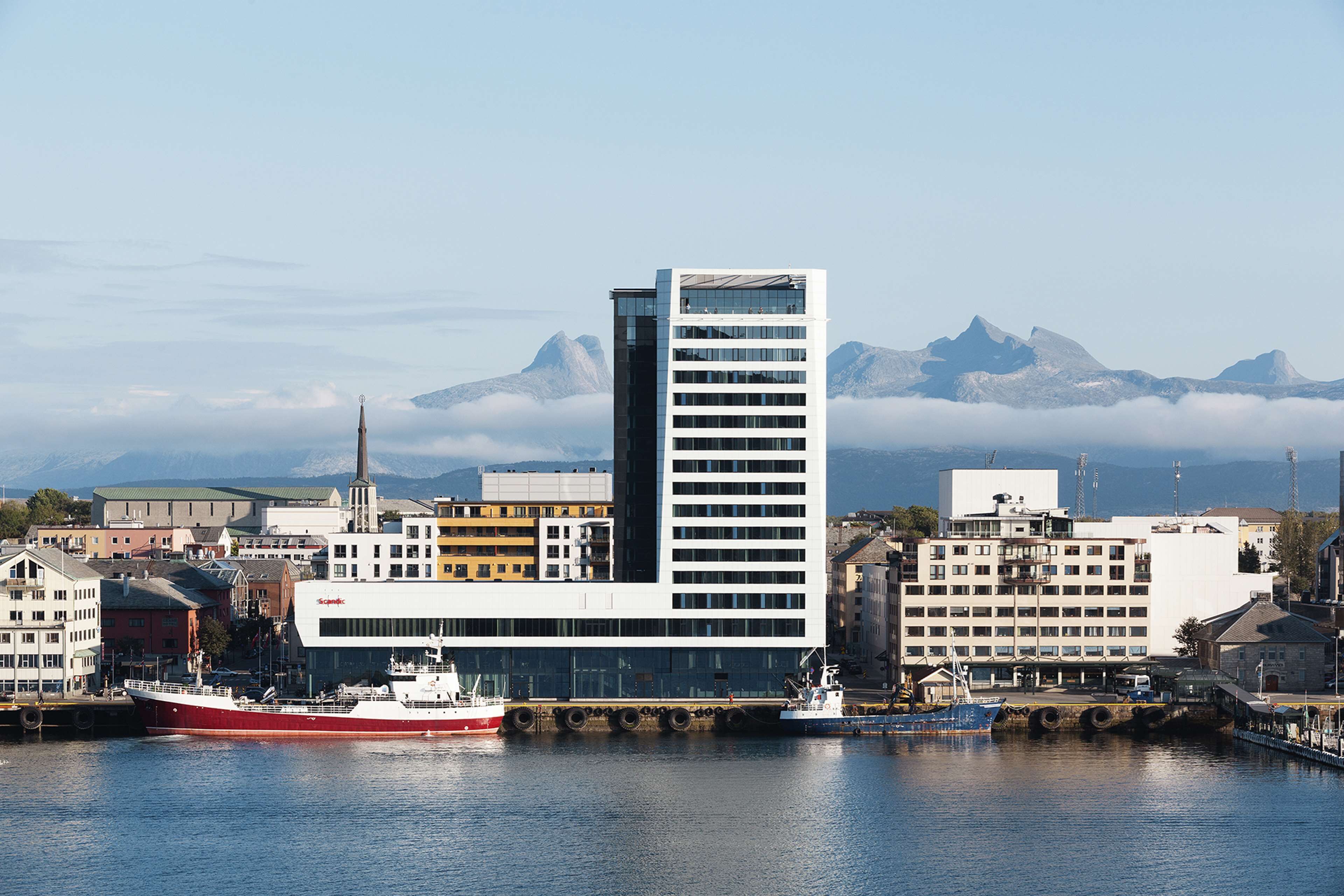 A modern building with a prominent tower, situated by a calm harbor with boats.