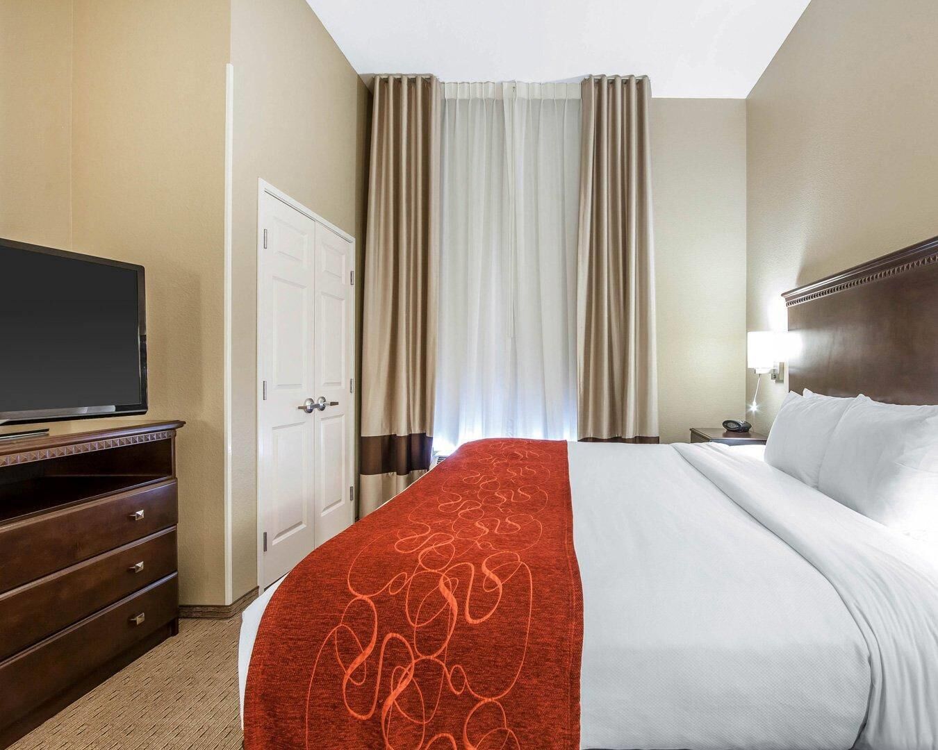 Habitación de hotel con una cama, televisor sobre un soporte, cómoda, puerta y cortinas.