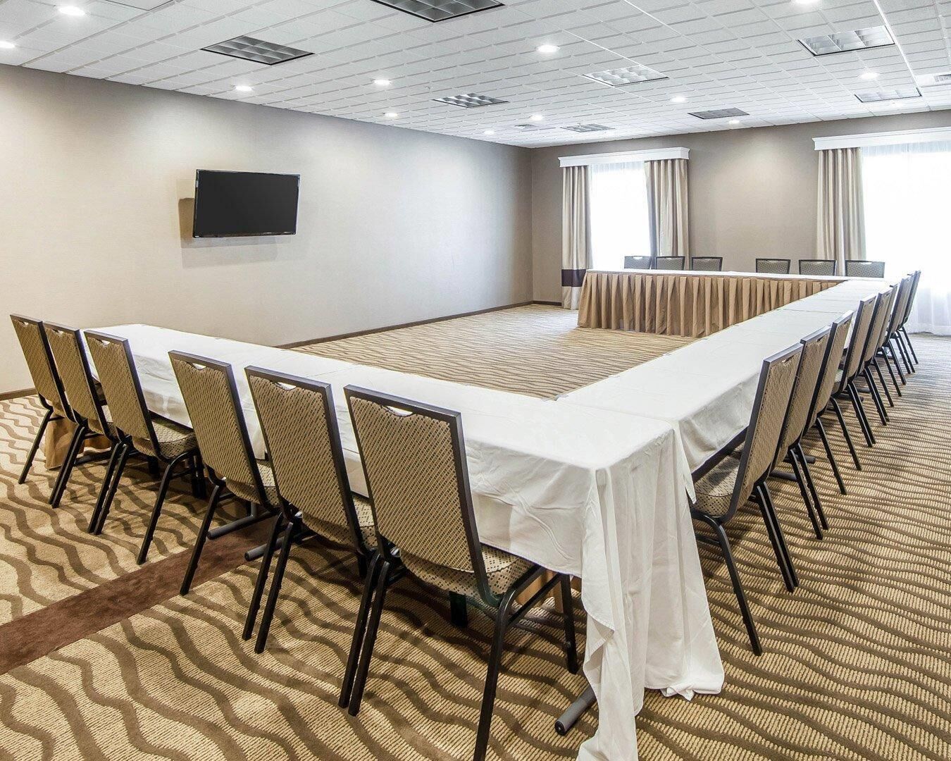 Un salón de conferencias con una larga mesa, sillas, un televisor y ventanas con cortinas.