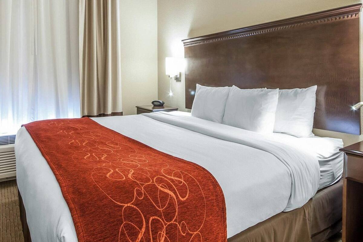 A hotel room with a large bed, a wooden headboard, a nightstand with a lamp, and a window with curtains.