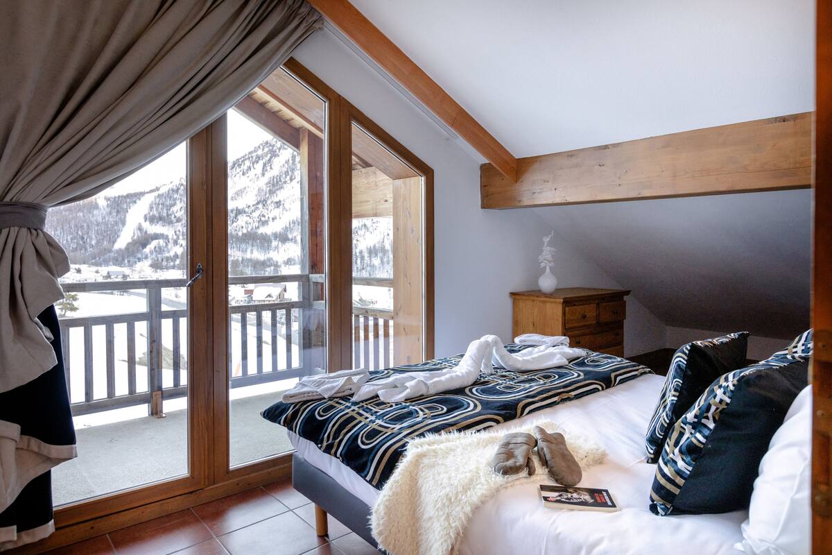 Une chambre avec une grande fenêtre, des poutres en bois, un lit avec une literie à motifs, un plaid blanc moelleux et une petite table de chevet en bois.