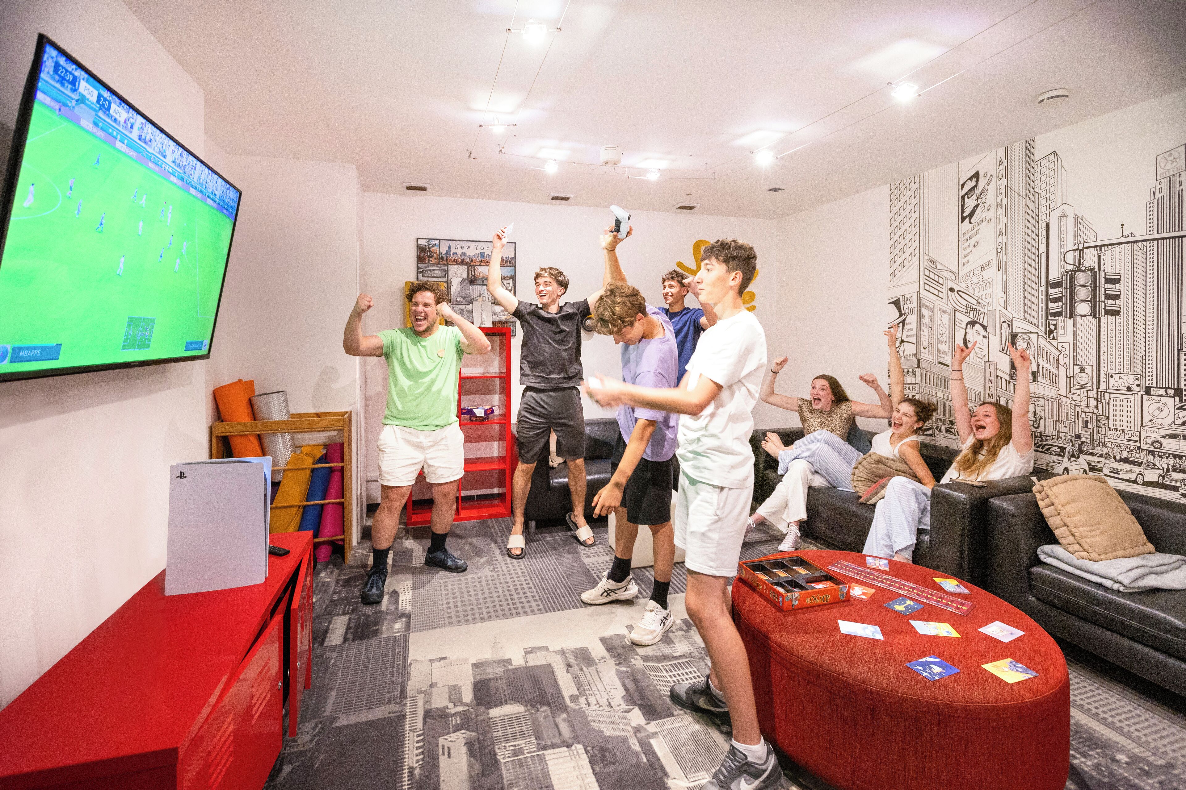 Een groep mensen speelt een videospel in een kamer met een grote televisie, een rode bureau en een muurschildering van een stadsgezicht.