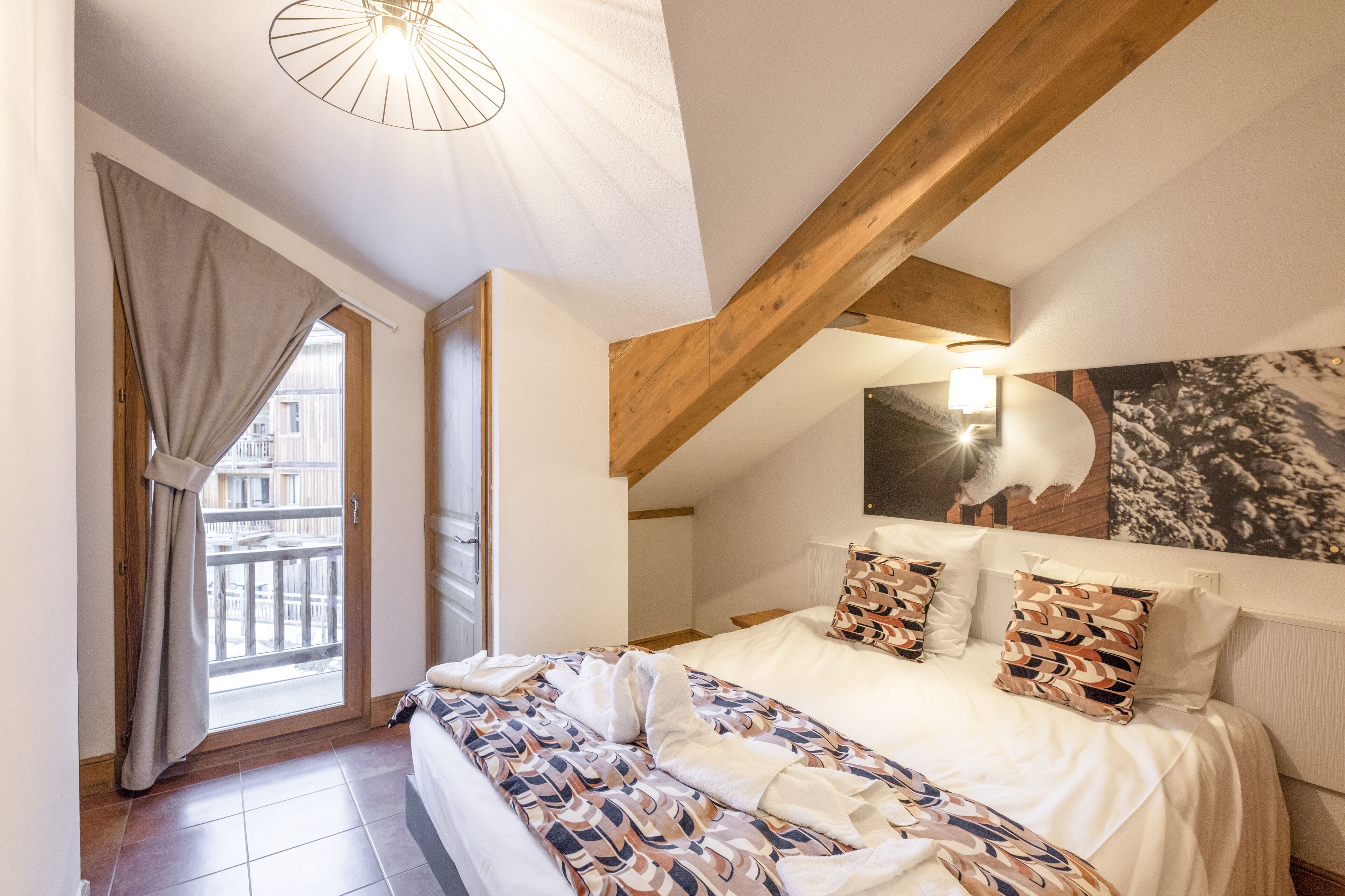 A bedroom with a large bed, wooden beams, a window with curtains, and a wall-mounted light fixture.