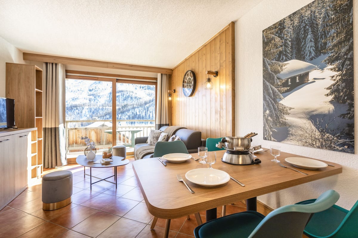 Un espace repas moderne avec une table en bois dressée pour deux personnes, un téléviseur en arrière-plan et une vue panoramique par la fenêtre.