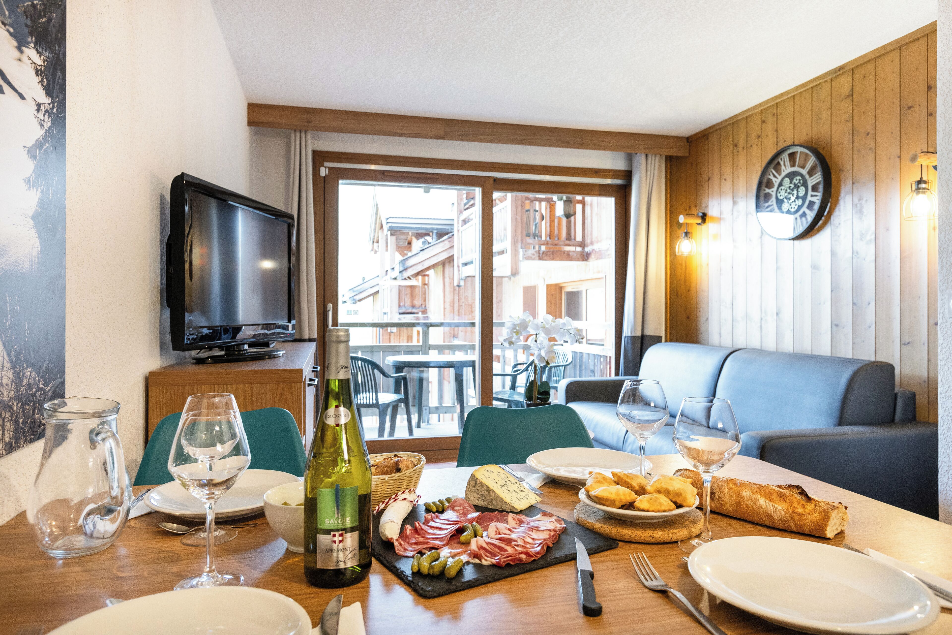 A dining area with a table set for two, featuring a bottle of wine, a cheese board, and a variety of meats.