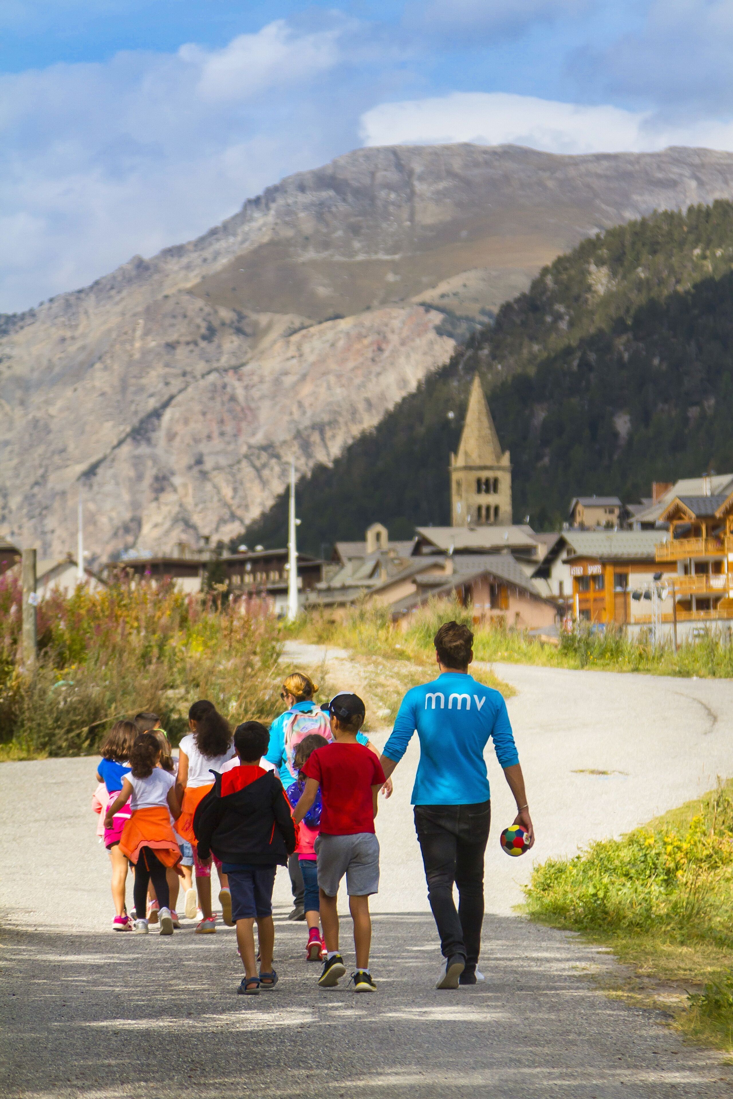 A group of people walking on a paved path with a mountainous backdrop and traditional buildings.