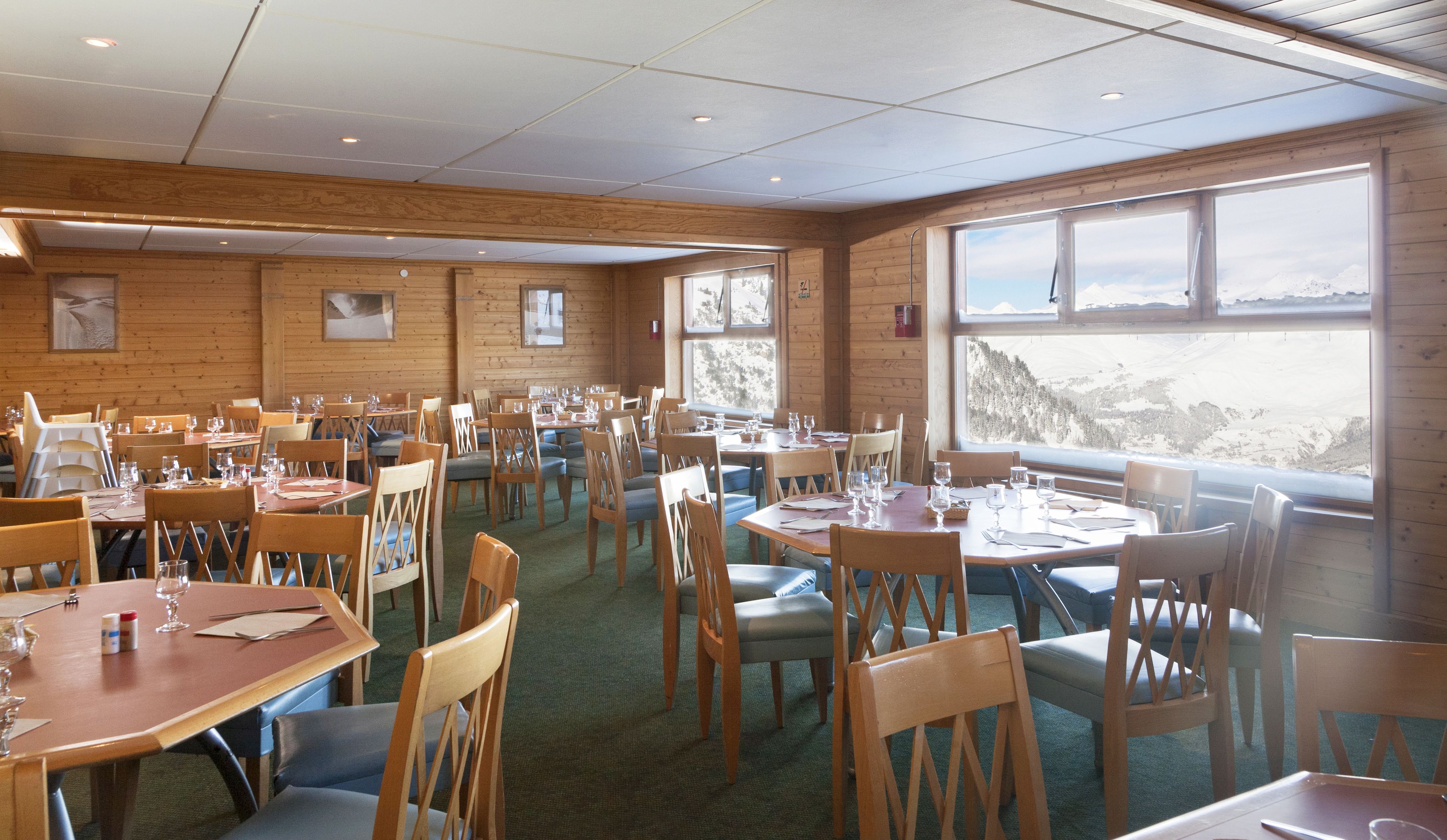 Un restaurant aux murs en bois et aux grandes fenêtres offrant une vue sur les montagnes. Les tables sont dressées avec des verres à vin et des assiettes.