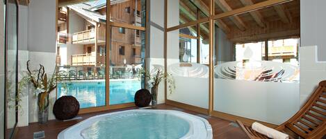 Zona de spa con un jacuzzi, sillones de madera y vistas a una piscina y edificios.