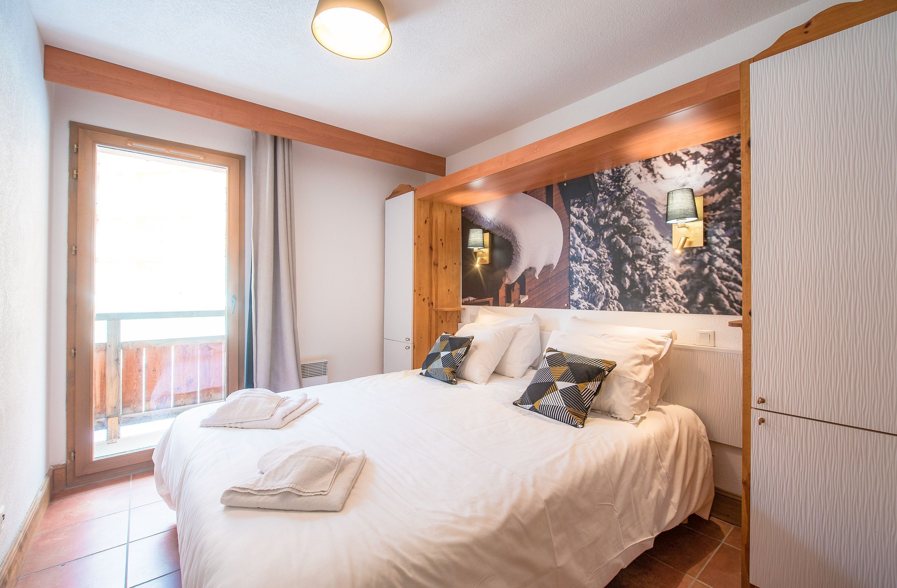 A bedroom with a large bed, a wooden ceiling, a window with curtains, and a wall-mounted lamp.