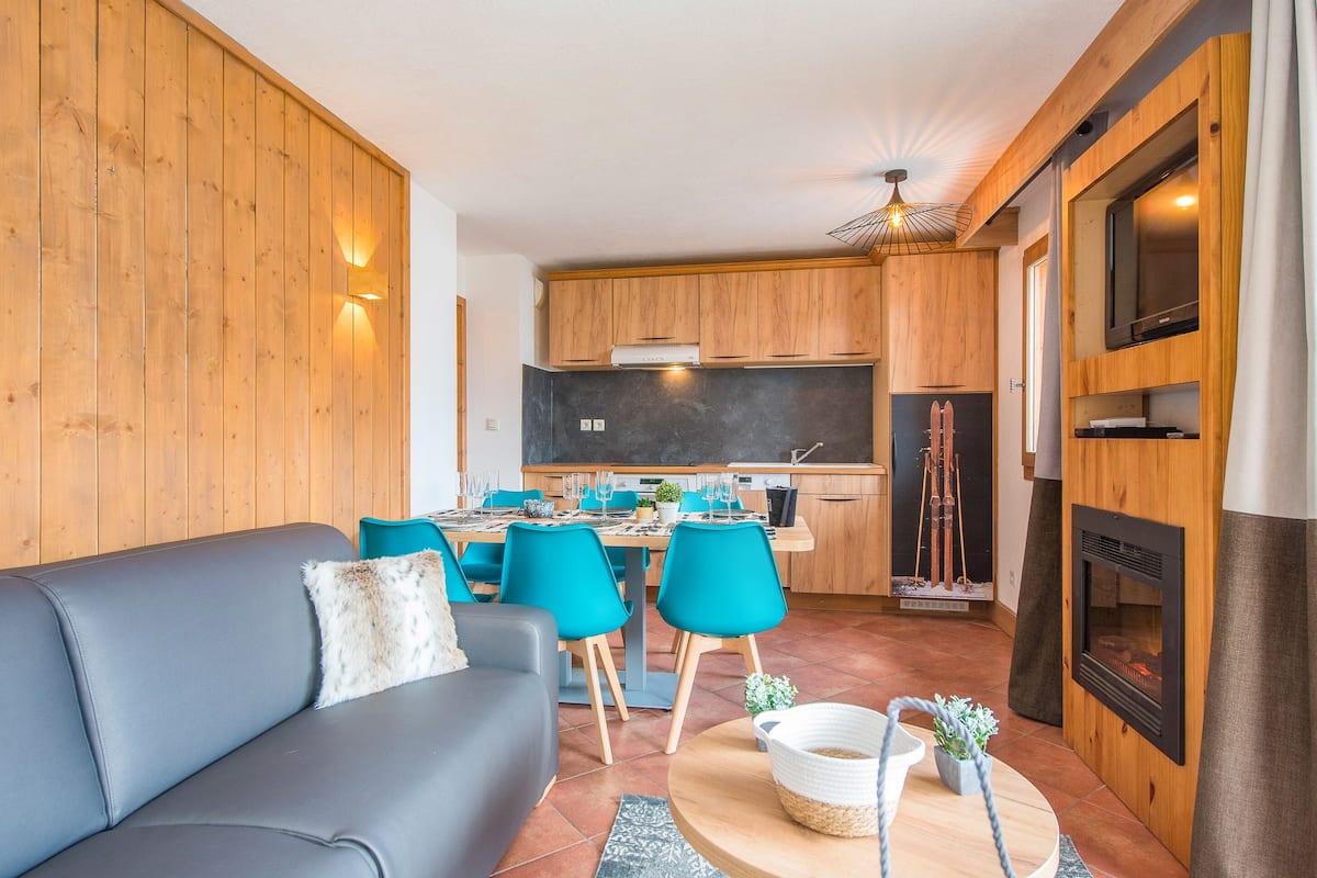 Un salon moderne avec un canapé gris, des fauteuils turquoise et une table à manger en bois, dressée pour six personnes.