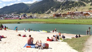 Una playa con gente tomando el sol y nadando, un lago y montañas al fondo.