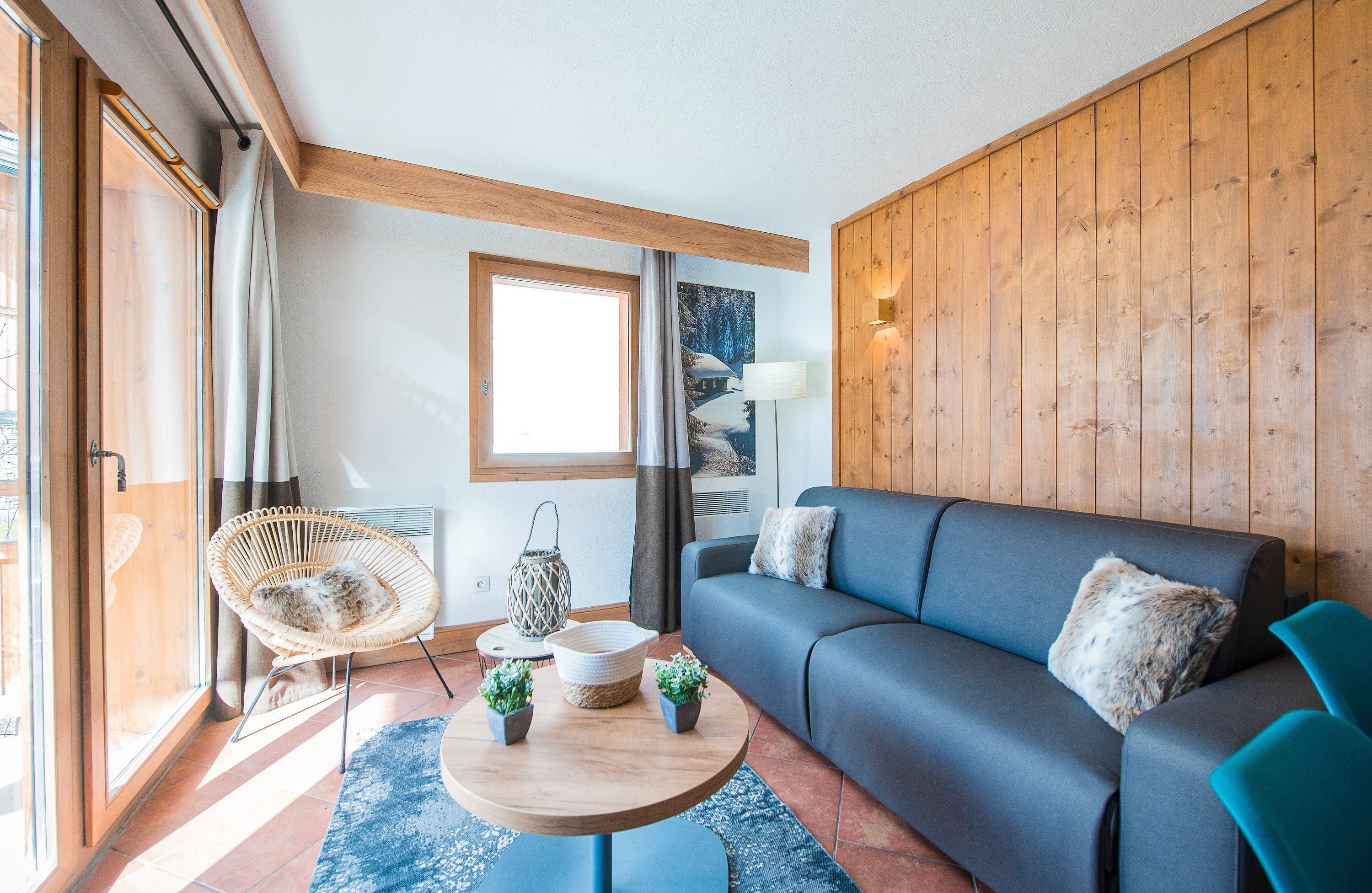 A cozy living room with a blue sofa, a wicker chair, a round wooden table, and a large window with curtains.