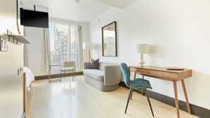 Une chambre d’hôtel moderne dotée d’un téléviseur à écran plat fixé au mur, d’un canapé gris, d’un bureau en bois avec une chaise et d’une grande fenêtre offrant une vue sur la ville.