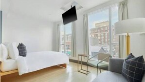 Une chambre d’hôtel moderne équipée d’un lit, d’un canapé, d’un téléviseur fixé au mur et de grandes fenêtres donnant sur la ville.