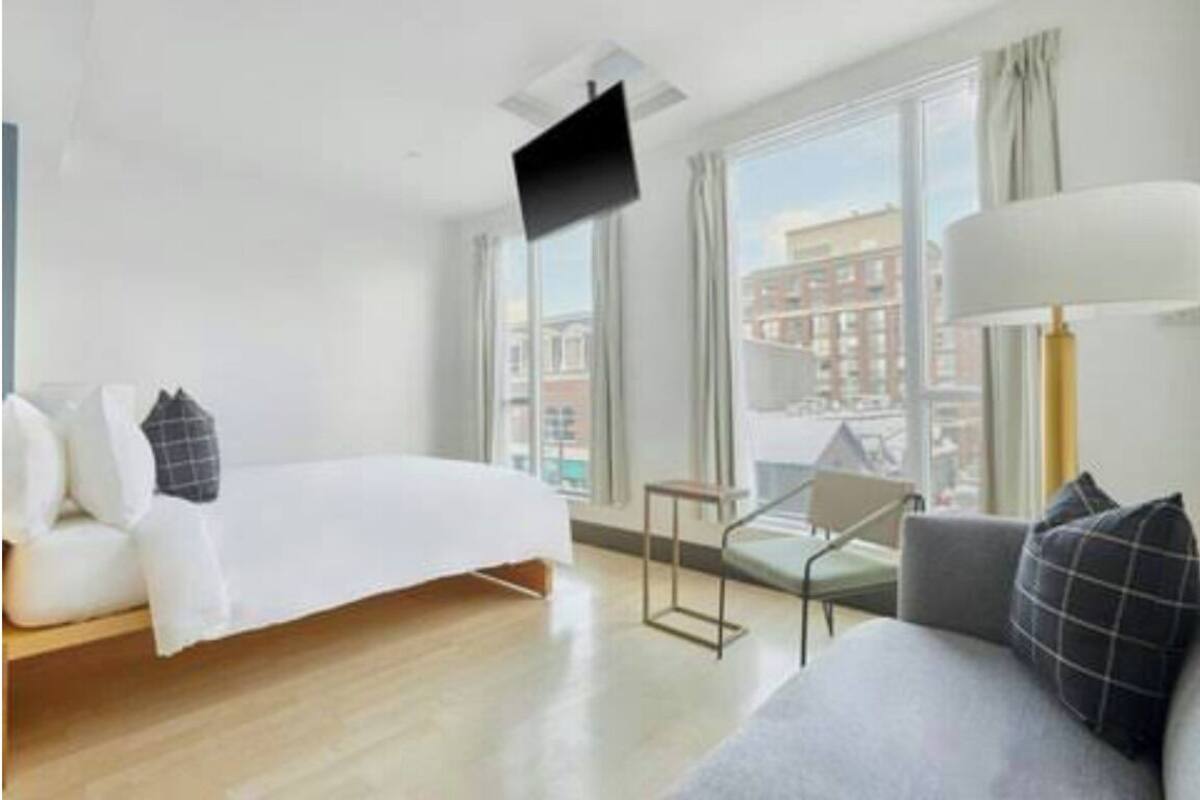 Una habitación de hotel moderna con cama, sofá, televisor montado en la pared y ventanas amplias con vistas a la ciudad.