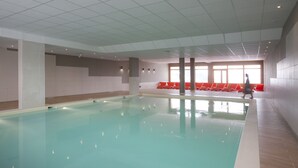Piscina interna com água cristalina, cadeiras de descanso vermelhas e uma grande janela que permite a entrada de luz natural.