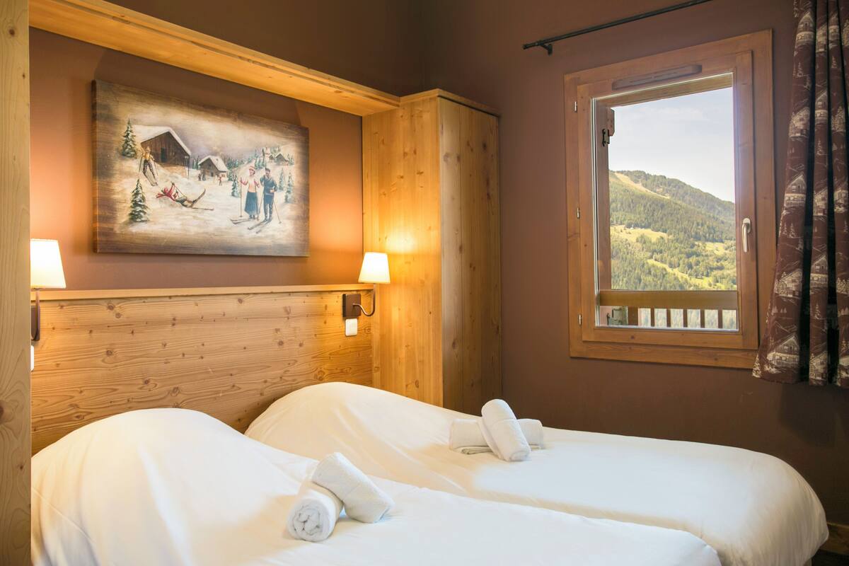A hotel room with two beds, a painting of a snowy scene, a window with a view of a forested mountain, and a wooden headboard.