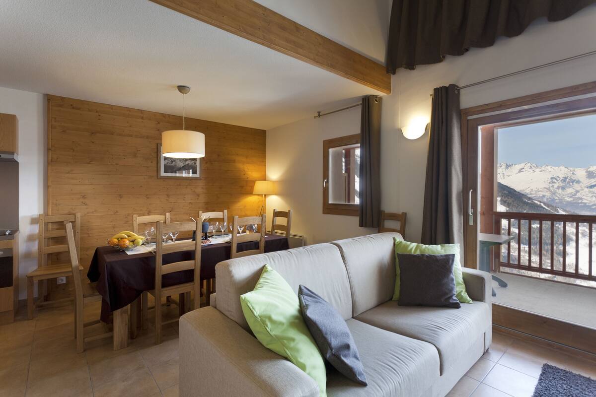 A cozy living room with a sofa, dining table, and chairs. A large window offers a mountain view.