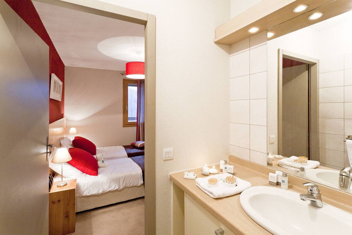 A hotel room with a bed, bedside table, lamp, and a bathroom with a sink, mirror, and toiletries.