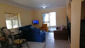 Un salon spacieux avec un canapé bleu, un fauteuil orange et un canapé marron. Un téléviseur est fixé au mur, et on aperçoit un escalier au fond de la pièce.