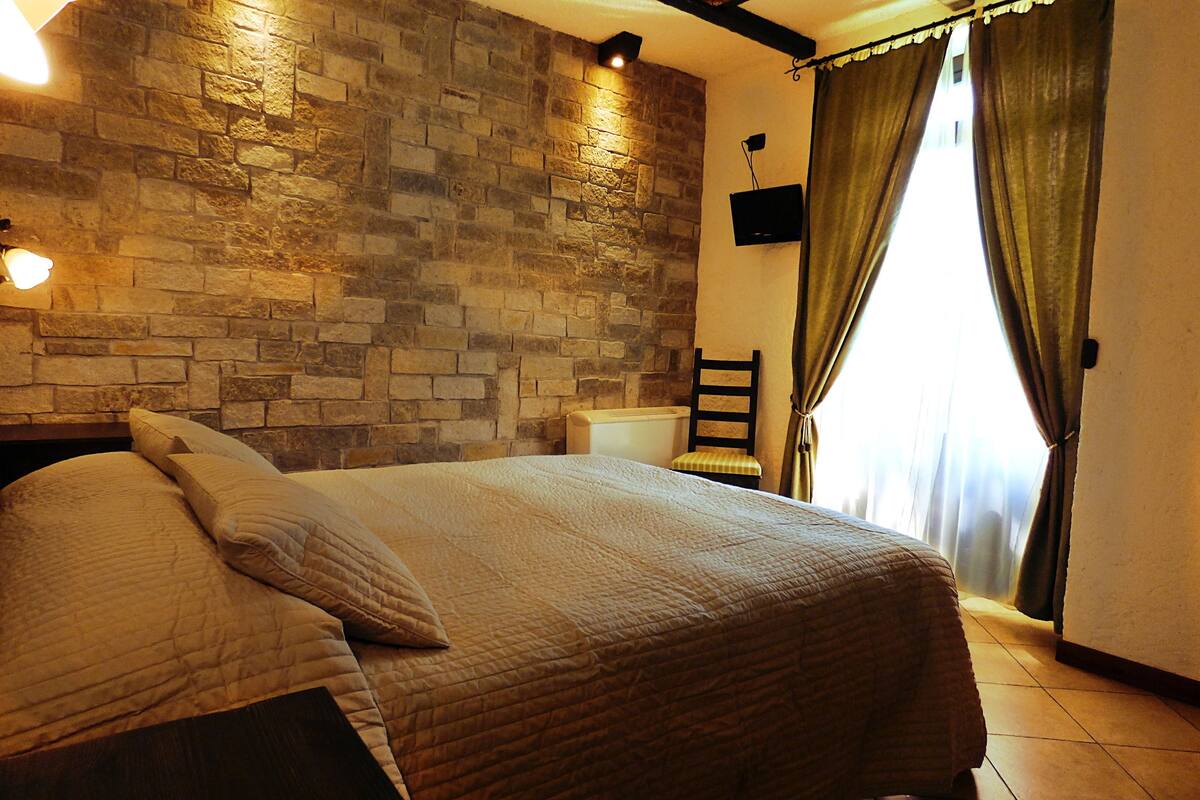 A bedroom with a stone wall, a bed with a quilt, a chair, a television, and a window with curtains.