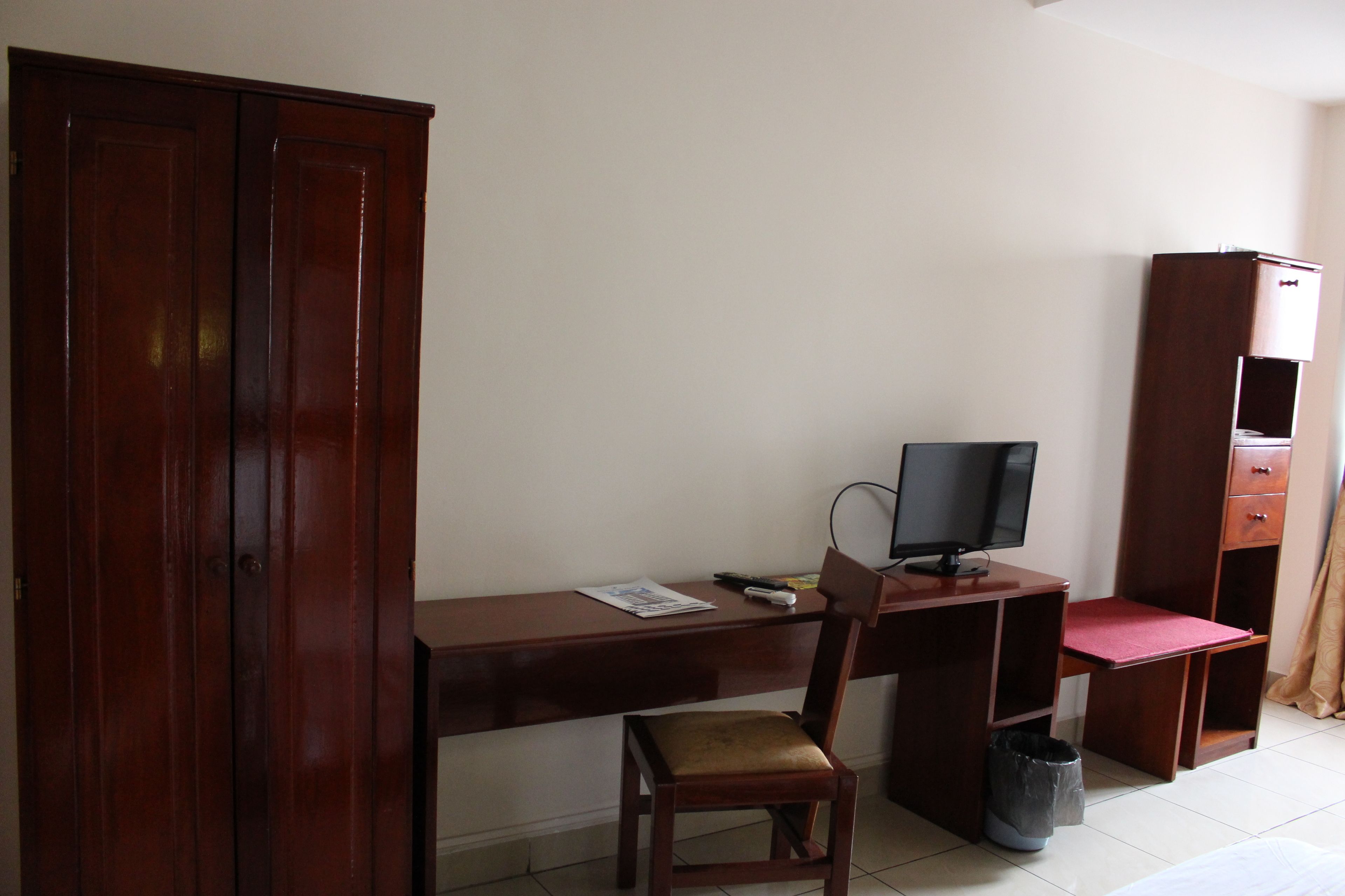 A room with a wooden desk, a chair, a television, a wardrobe, and a small trash bin.